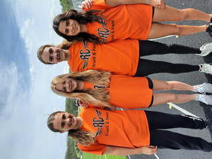 four female track athletes stand smiling on a track wearing orange RC shirts