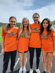 four female track athletes stand smiling on a track wearing orange RC shirts